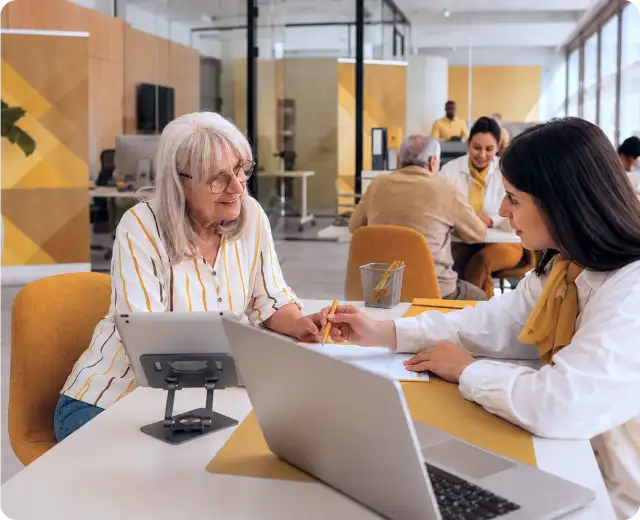 A bank representative showing information on a tablet and laptop to an elderly woman in a busy, modern branch.